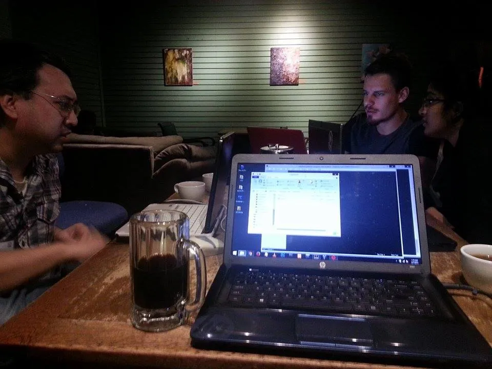 Three of us around a table in a dim coffeeshop, two laptops open and a glass mug of black coffee in the foreground