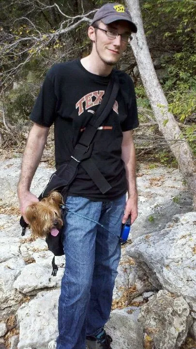 Me on a rocky trail in a Texas T-shirt and a baseball cap, a small terrier riding in a sling bag at my hip and a water bottle in my hand